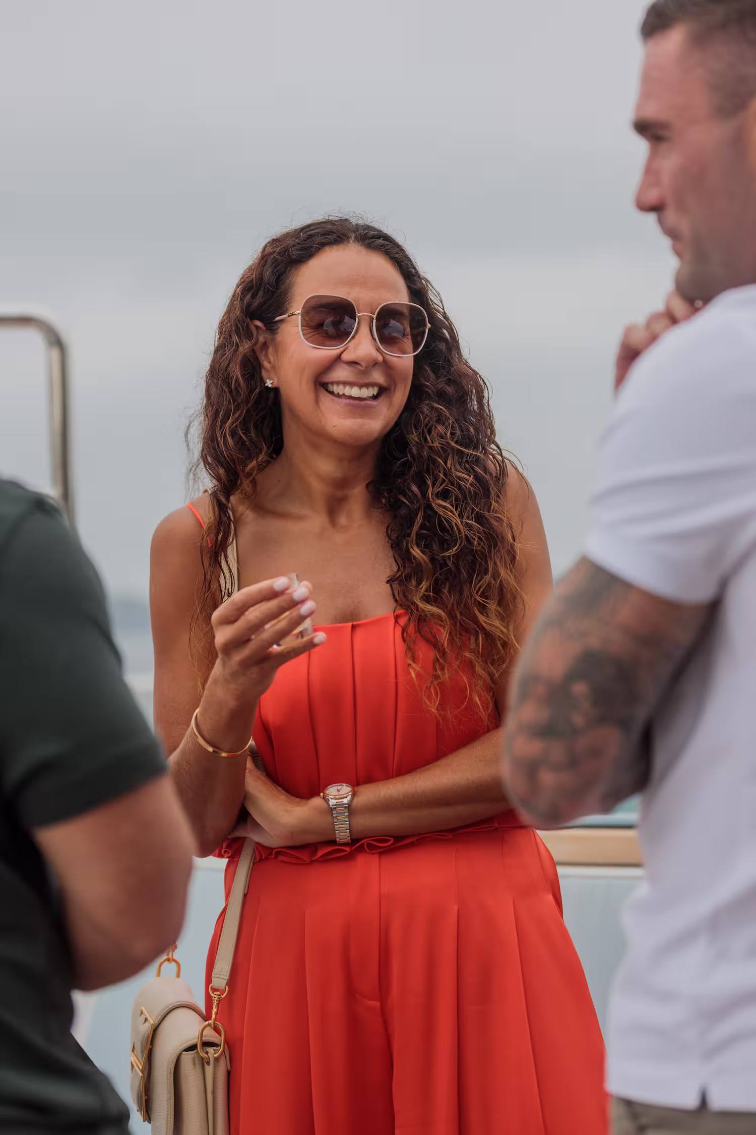 A woman wearing a red dress and sunglasses stands outdoors, smiling.