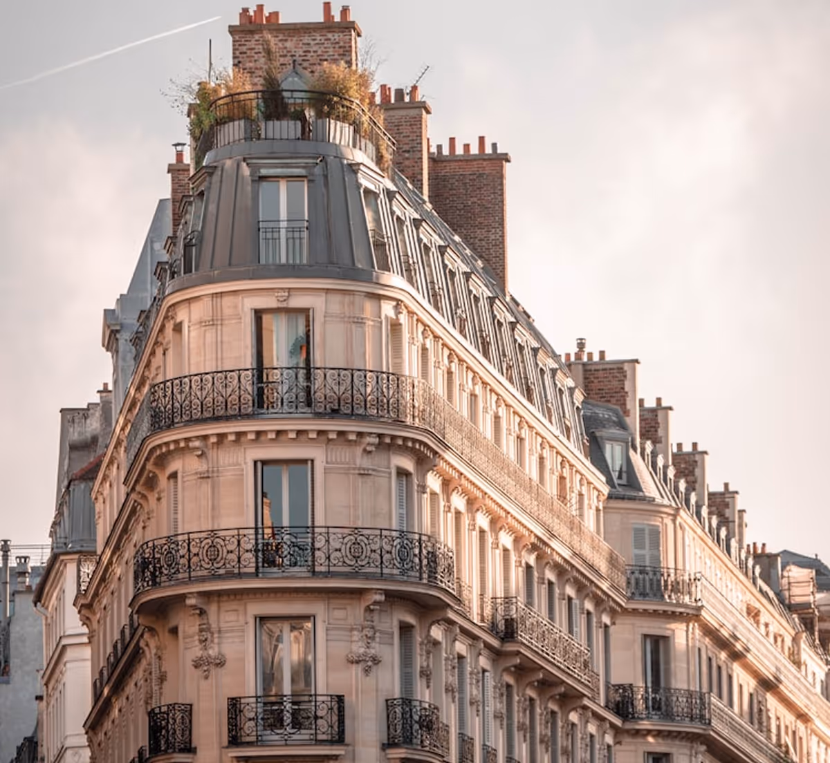 Immeuble haussmannien d'angle avec balcons en fer forgé et toit mansardé, baigné par une lumière chaude.