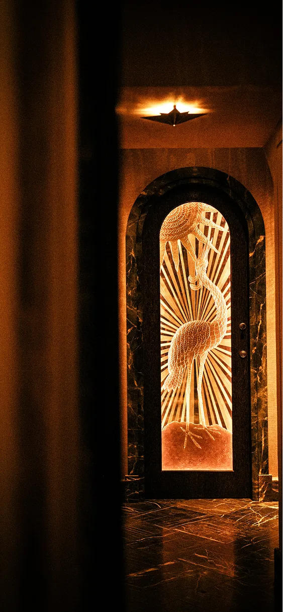 Porte avec un panneau en verre gravé d'un oiseau au long cou, éclairée d'une lumière chaude dans un intérieur sombre.