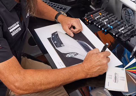Person sketching a futuristic car design on paper with markers and color samples nearby.