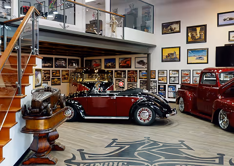 Vintage car showroom featuring a polished red and black convertible, a classic red truck, framed automotive artwork on walls, and a wooden staircase with glass railing.