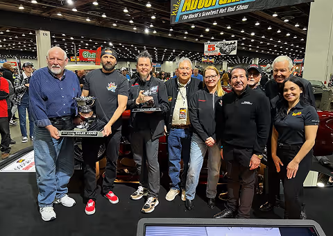 Group of eight people standing indoors at a car show, some holding trophies, surrounded by crowd and exhibition displays.