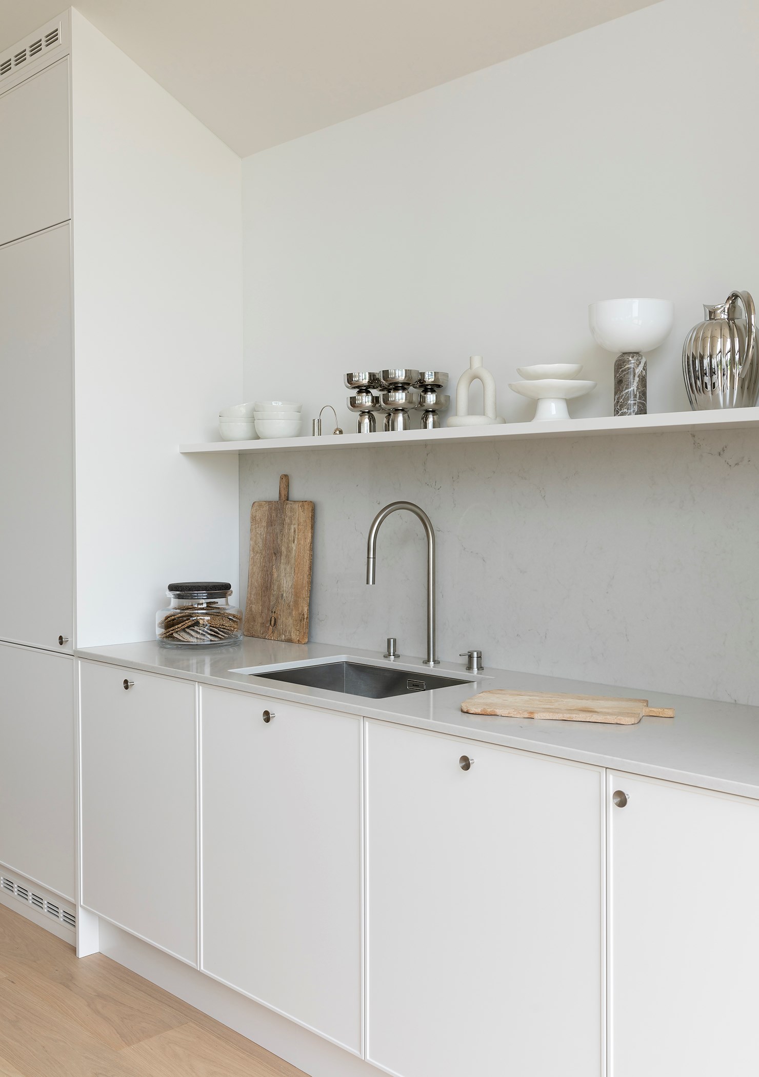 Minimalistisk hvitt kjøkken med marmorbakvegg, rustfri stålvask, åpen hylle med dekorative gjenstander og trefjøler.