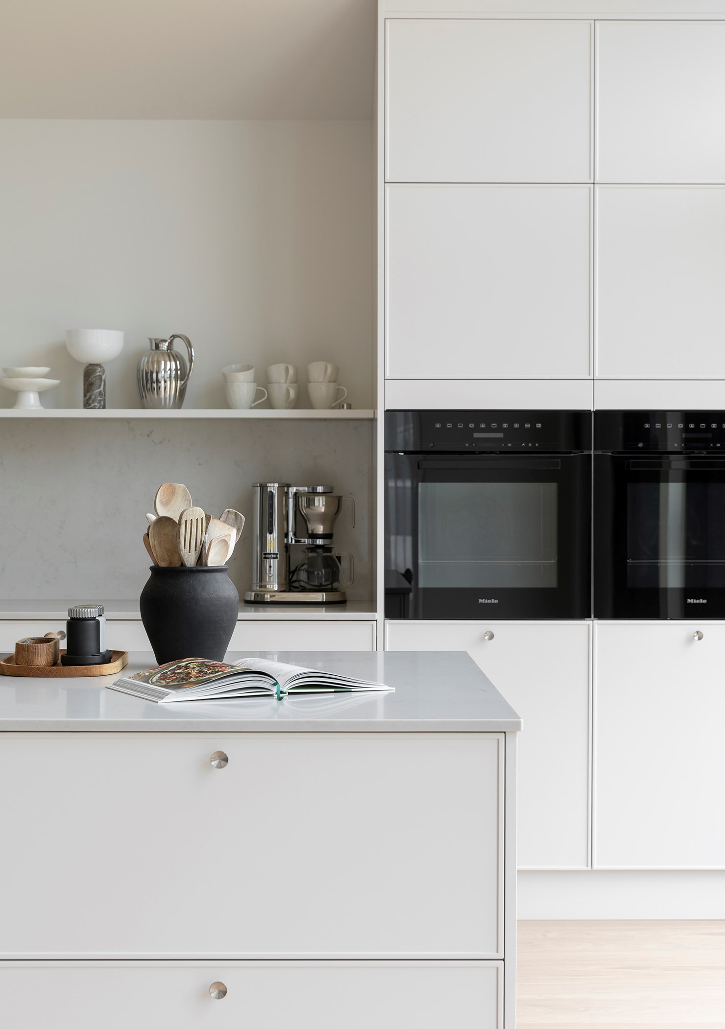 Moderne hvitt kjøkken med Miele-ovner, kjøkkenøy og åpen kokebok, samt hyller med kopper og kjøkkenutstyr.