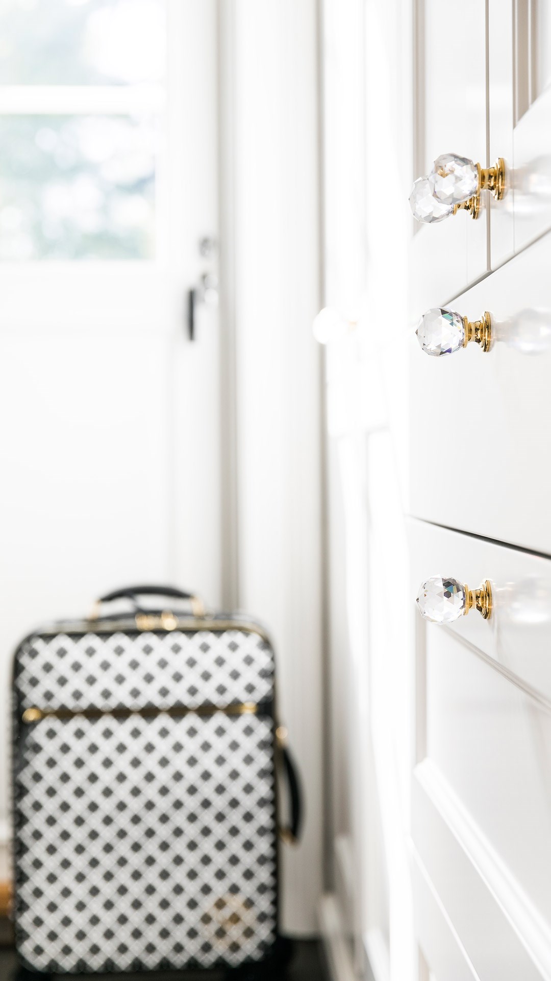 Hvit garderobe med krystallknotter i gull, og en svart-hvit rutete koffert i bakgrunnen.