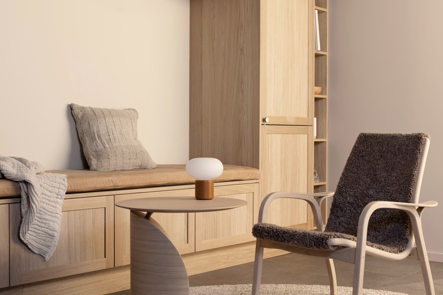 Minimalistisk stue med innebygd garderobe og benk i lyst tre, rund sofabord og stol med saueskinnstrekk.