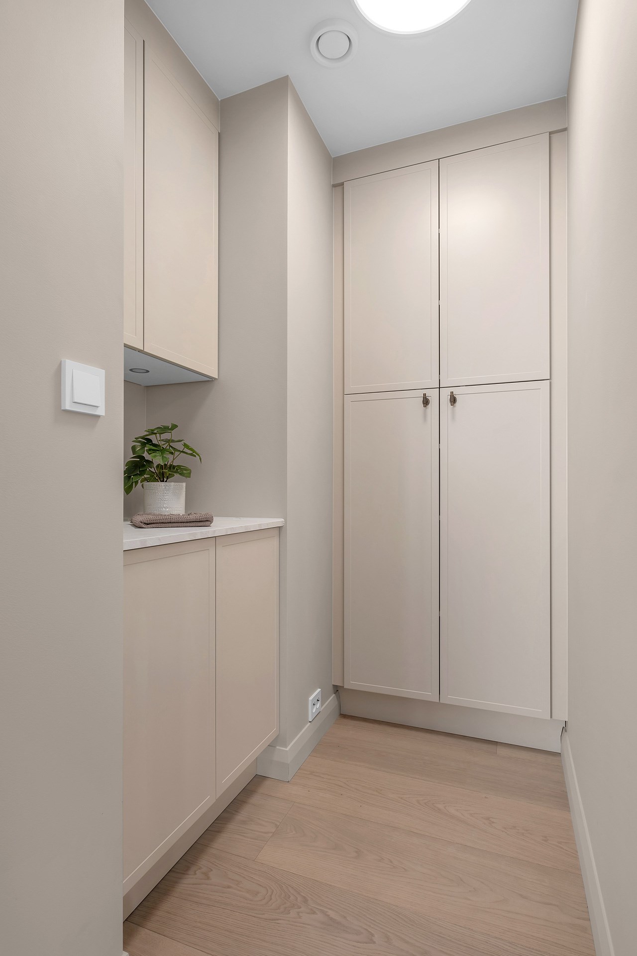 Smal garderobe med sandfarget innredning, høye skap og en liten hylle med grønn plante.