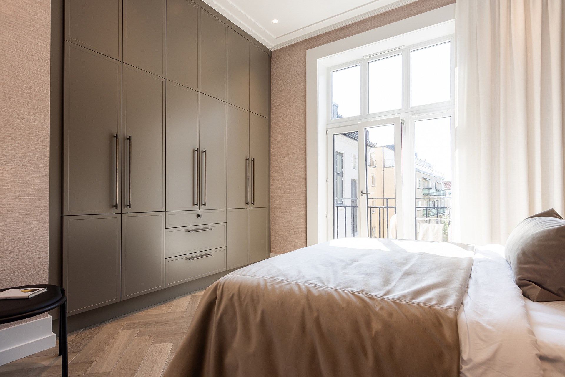 Soverom med stor innebygget garderobe i gråbrun farge, dobbeltseng og stort vindu mot balkong.