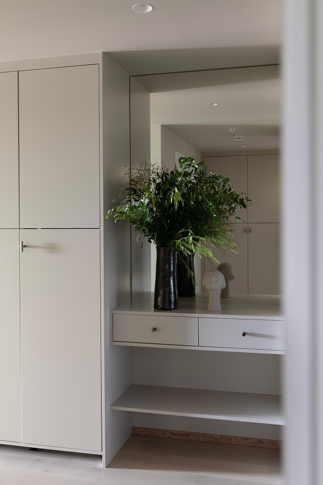 Innebygd garderobe i lys grå med speil, flytende skuff-konsoll og mørk vase med grønne planter.