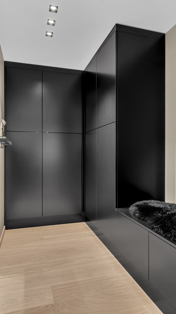 Moderne svart garderobe med innebygde skap og benk med mørk pelsdetalj, lyst tregulv og innfelte spotlights i taket.