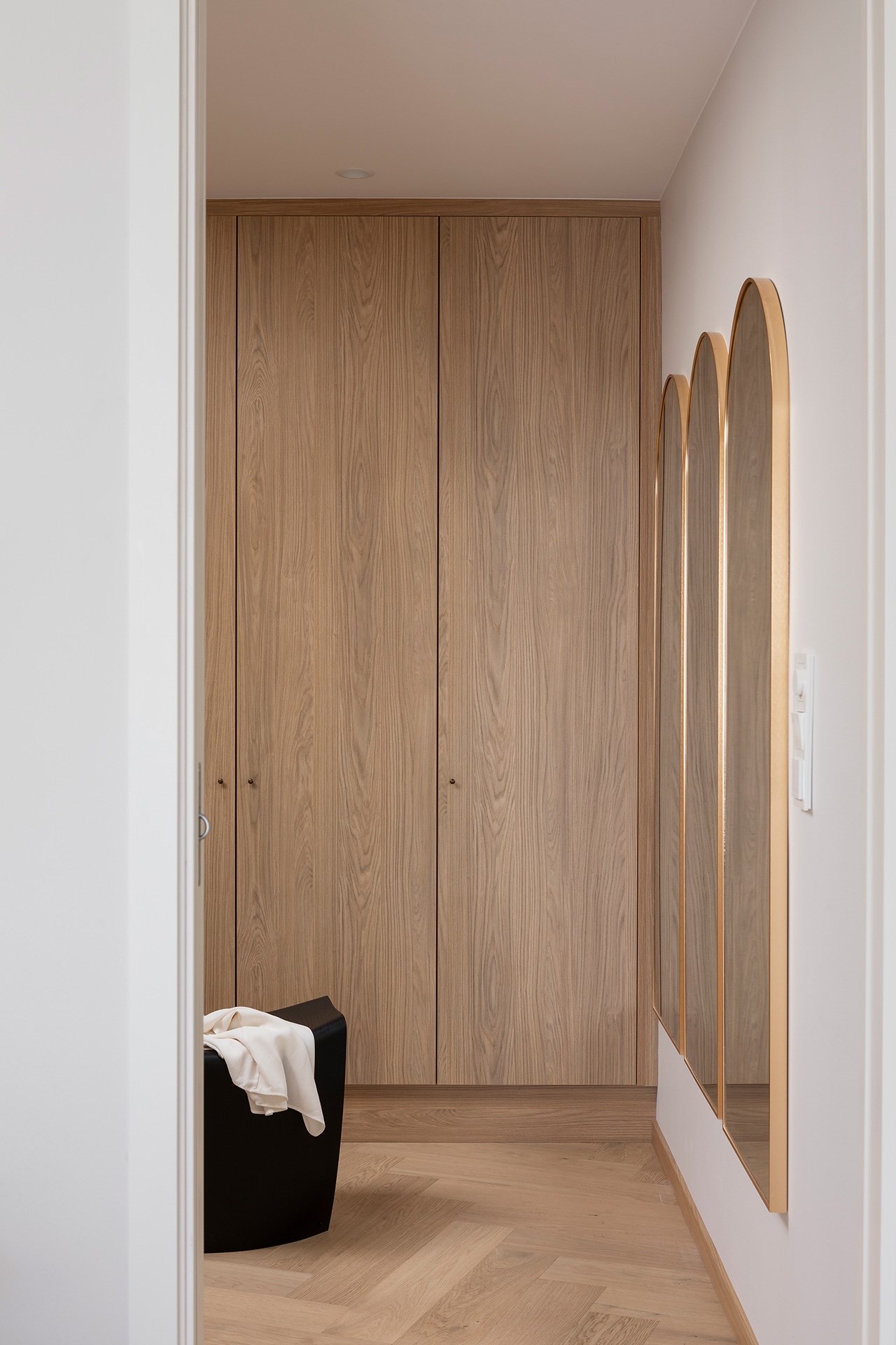 Minimalistisk garderobe med eikefronter, chevrongulv og buede speil med messingkant på hvit vegg.