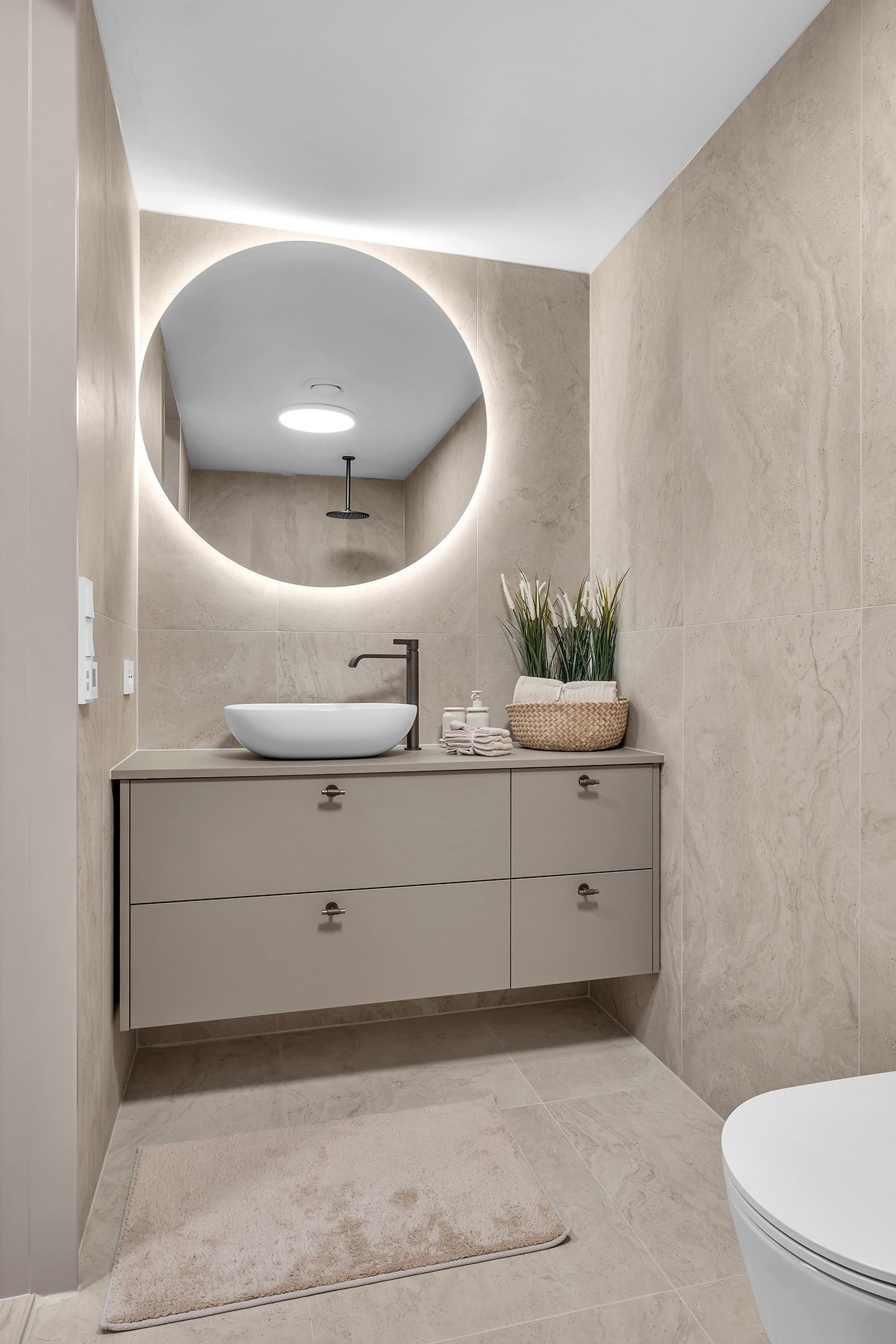 Moderne bad med flytende beige baderomsinnredning, rundt speil med bakbelysning og hvit servant.
