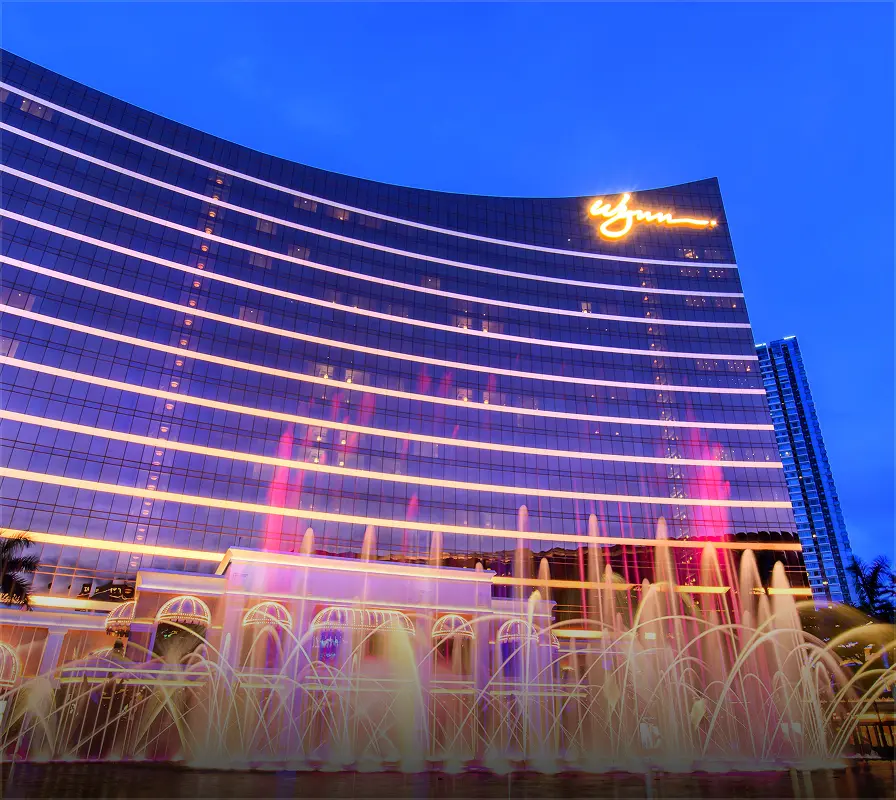 Wynn hotel building at dusk with illuminated horizontal lines and colorful fountains in front.