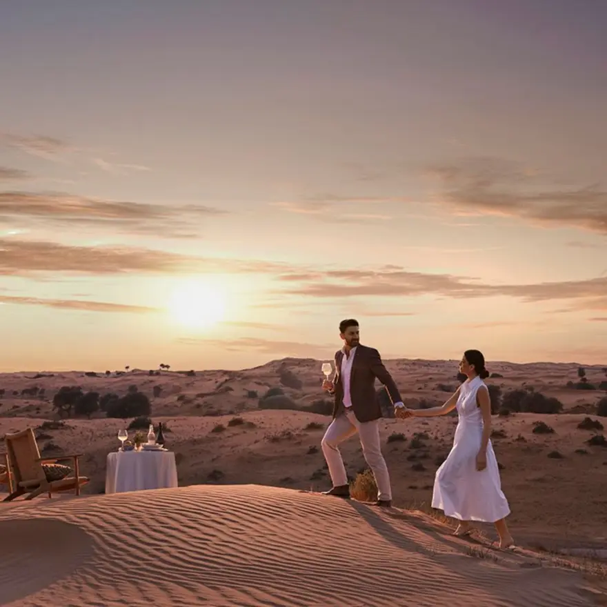 Couple dressed elegantly, holding hands while walking on sand dunes at sunset with a table set for two nearby.
