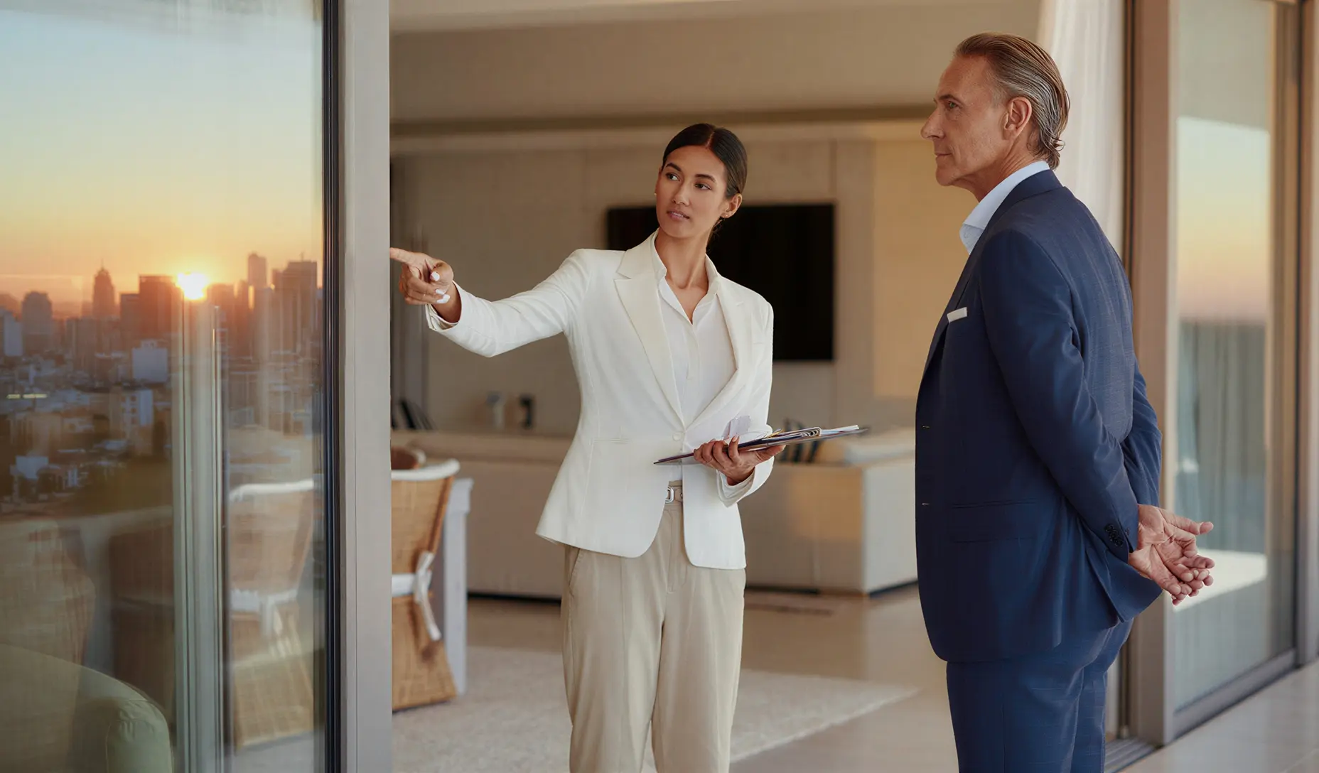 Real estate agent showing a property to a man by a large window with a city skyline at sunset visible outside.