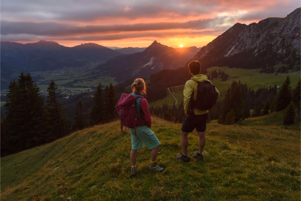 Zwei Wanderer mit Rucksäcken stehen auf einem grasbewachsenen Hügel und schauen auf ein Tal mit Bergen bei Sonnenuntergang.