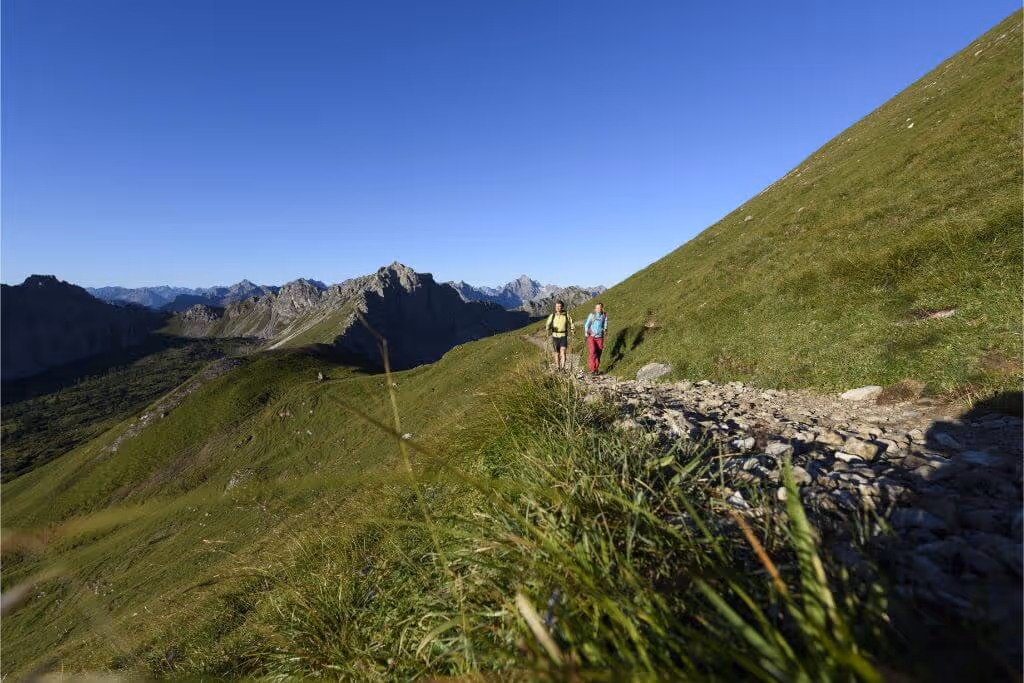 Zwei Wanderer auf einem felsigen Pfad in grünen Alpenwiesen mit Bergpanorama unter klarem blauem Himmel.