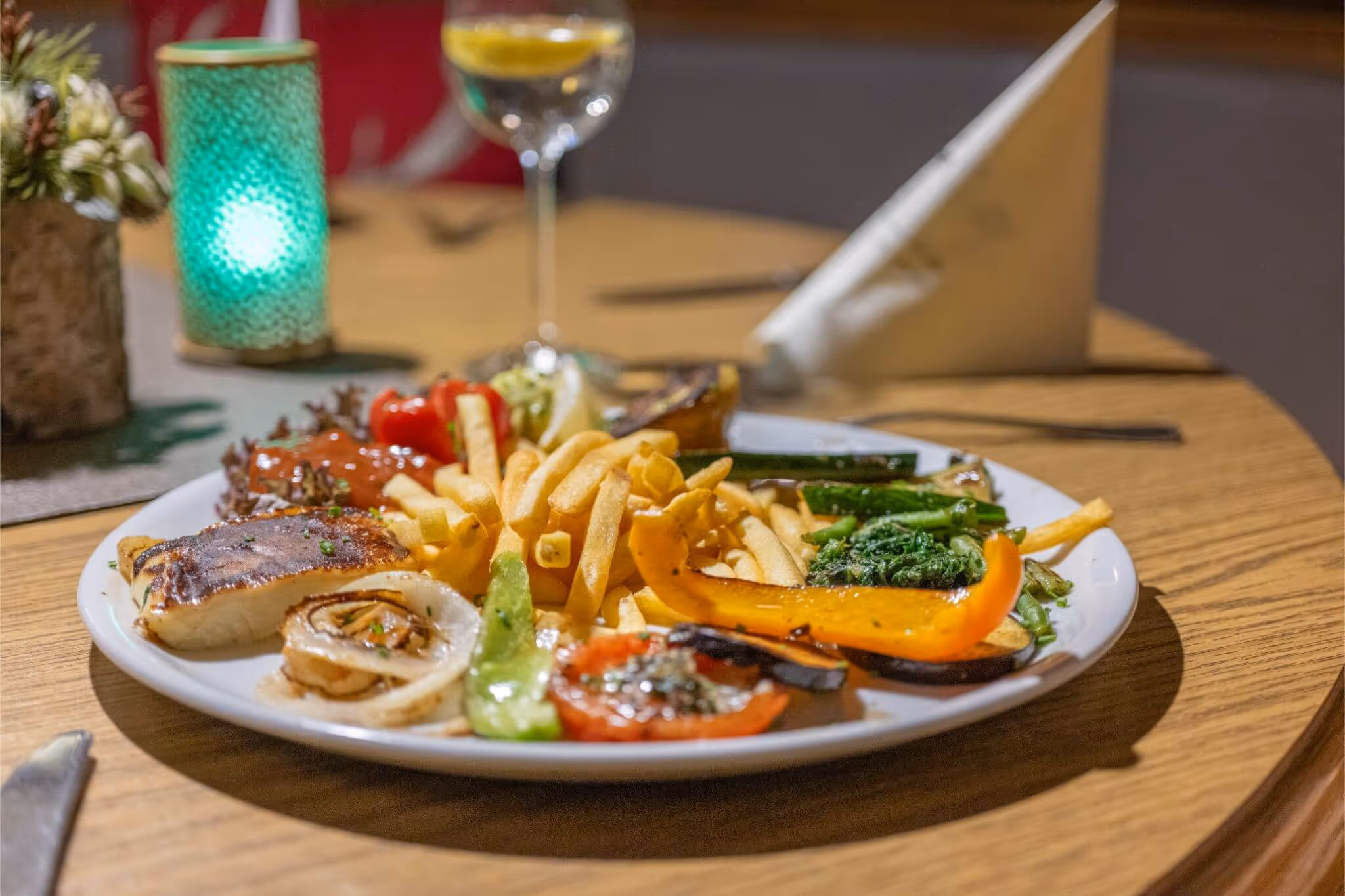 Teller mit gegrilltem Gemüse, Pommes frites, Salat und einem Getränk im Hintergrund auf einem Holztisch.