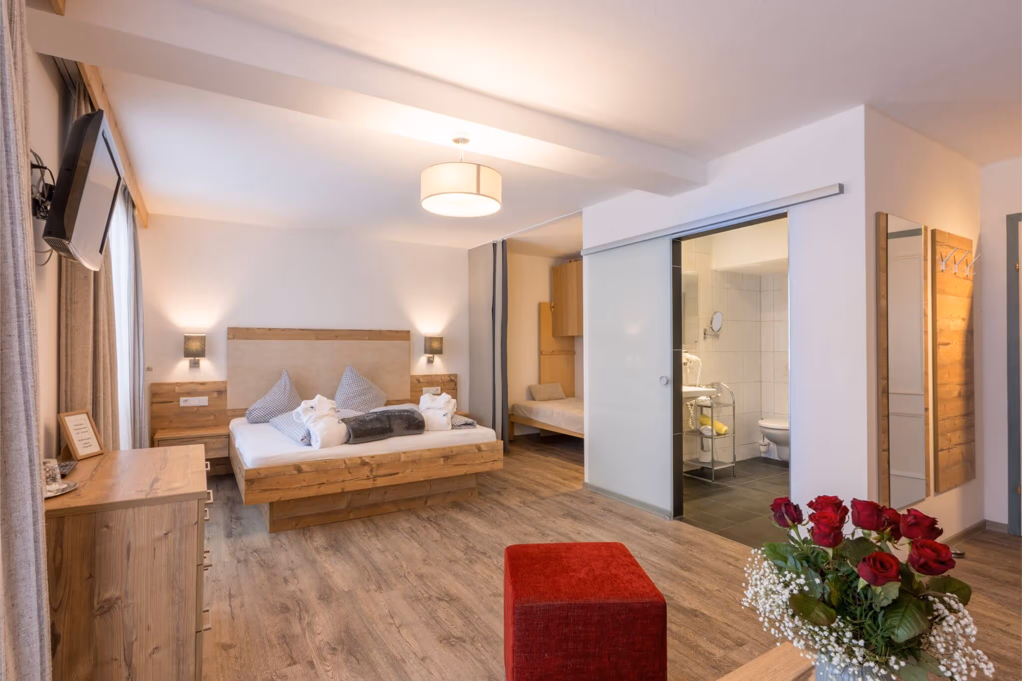 Modernes Familienzimmer mit Holzmöbeln, Doppelbett, Einzelbett, rotem Hocker und Blumenstrauß auf dem Tisch.