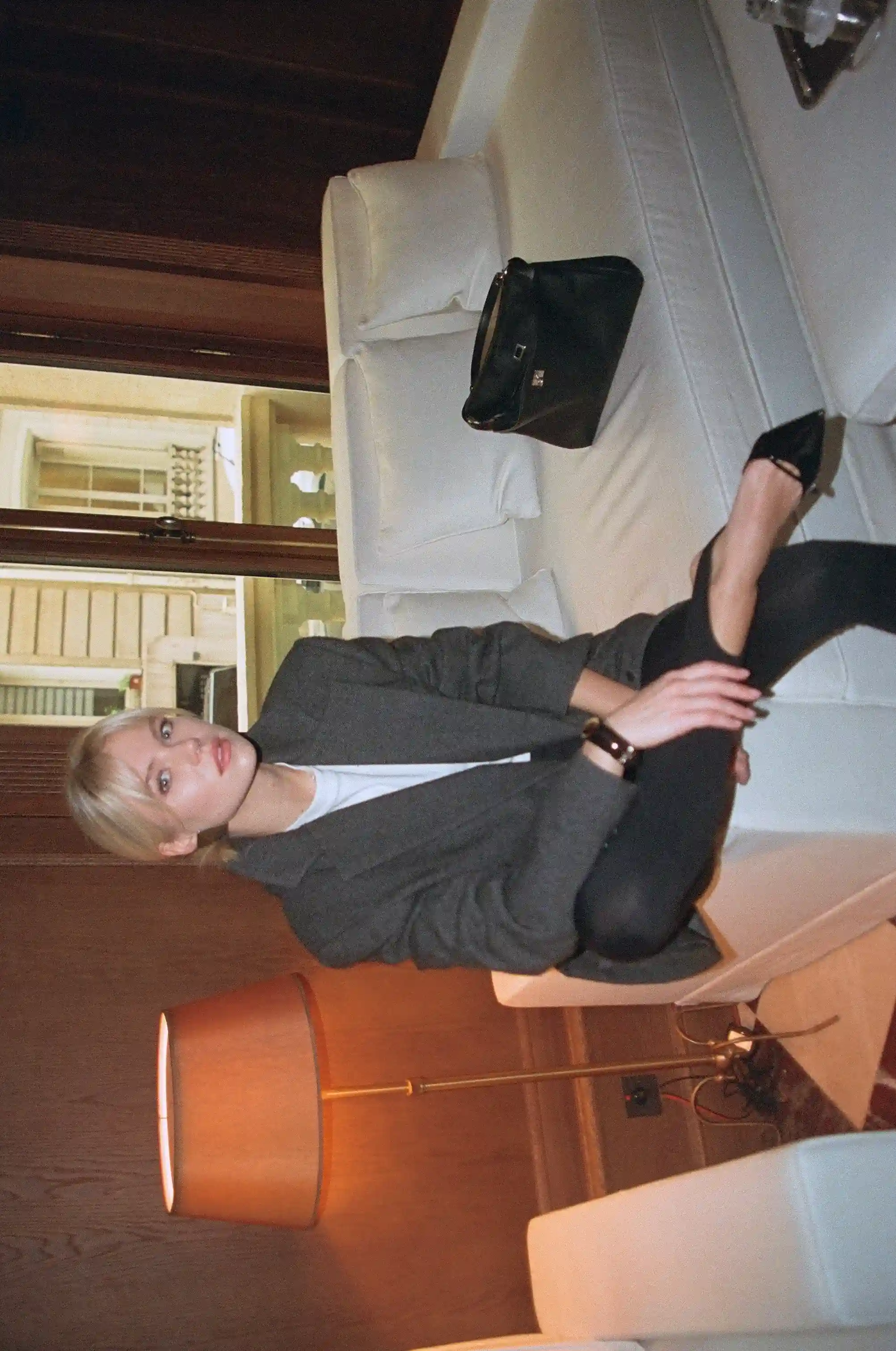 Woman with blonde hair wearing a gray blazer and black high heels sitting on a white couch in a room with wooden walls and a floor lamp.