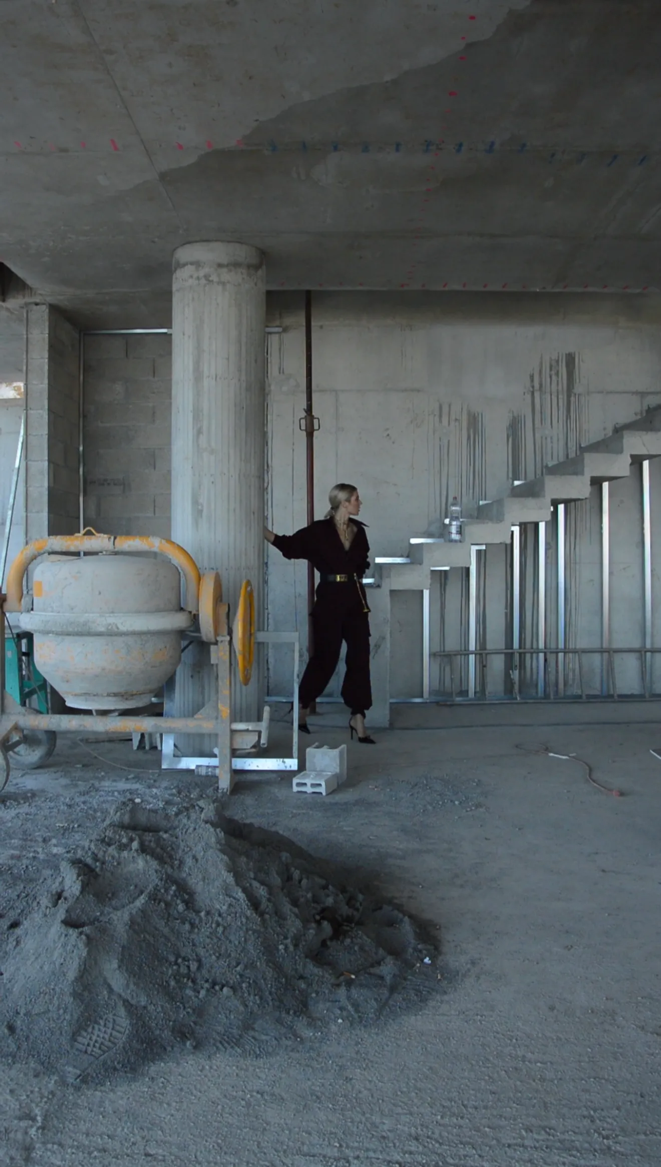 Person in dark clothing and heels standing in an unfinished concrete building near a cement mixer and stairs.