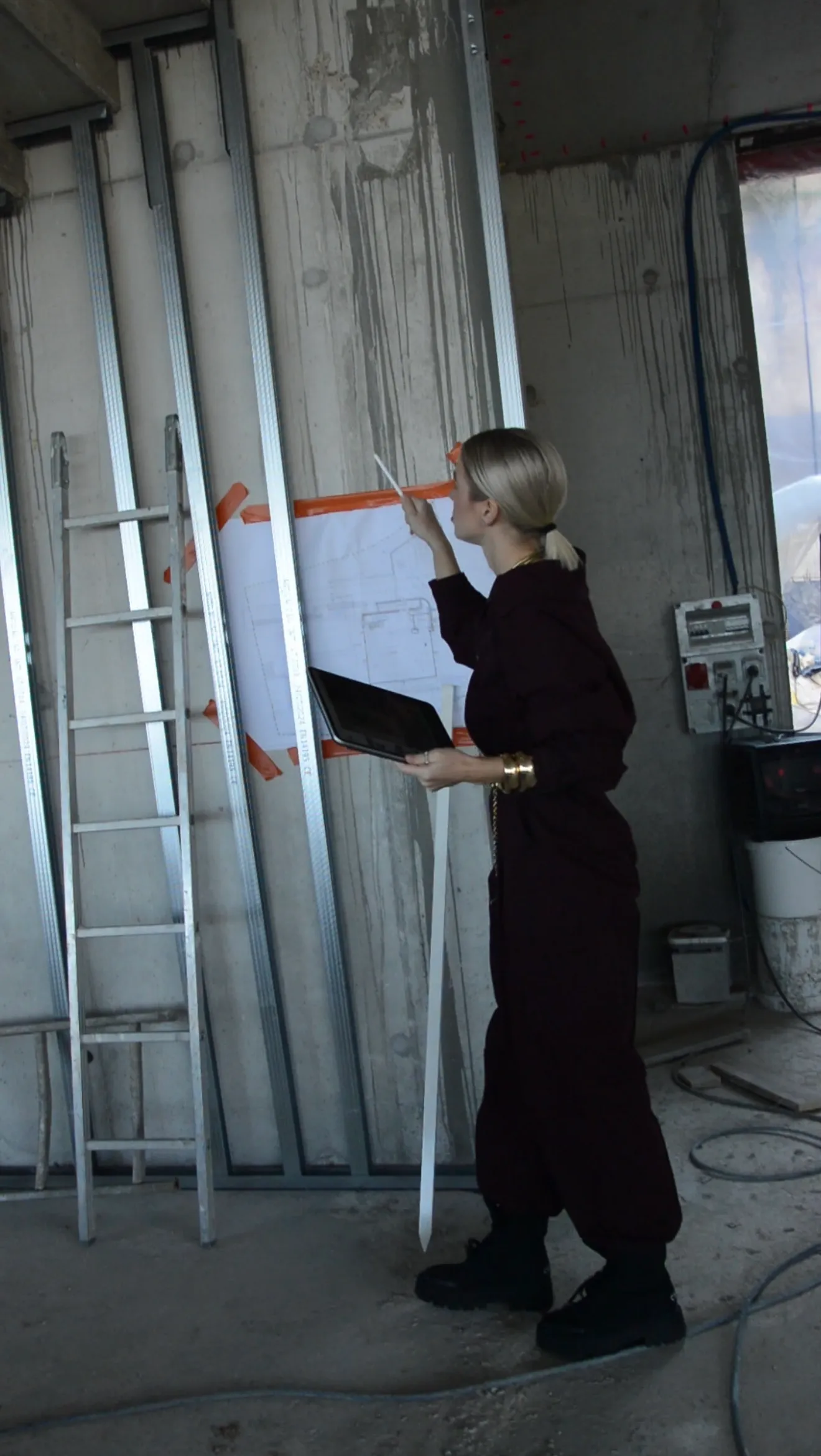 Woman in dark clothing holding a tablet and pointing at a building plan taped to a concrete wall in a construction site.