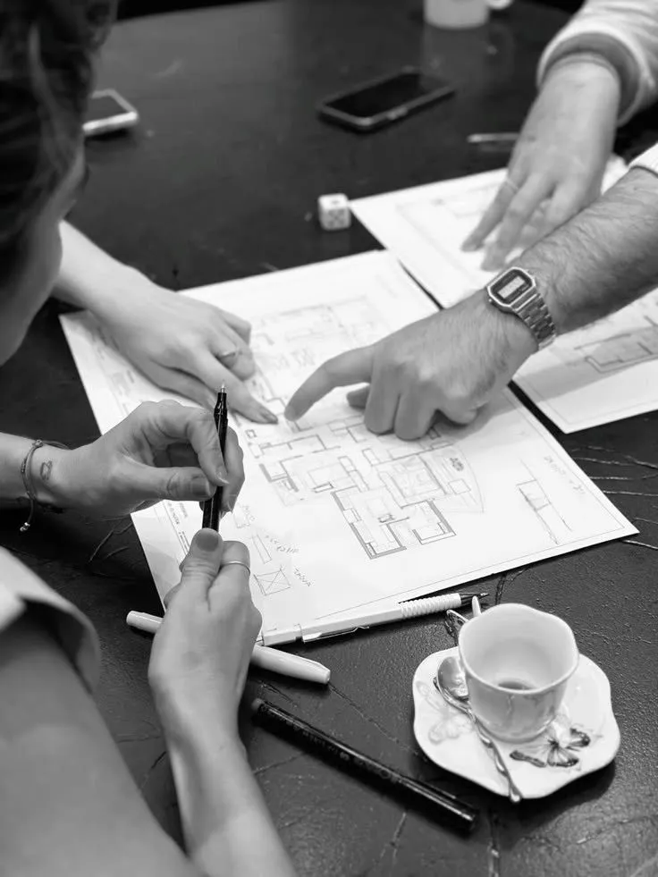 Two people reviewing architectural floor plans on a table with pens, a smartphone, a dice, and a partially filled coffee cup on a saucer.