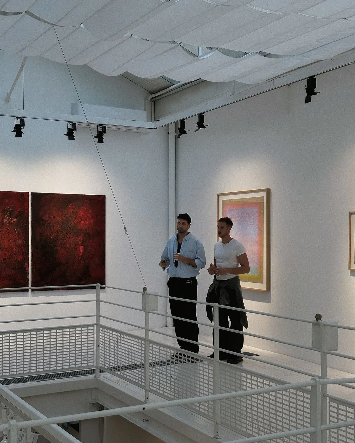 Two men standing on a white gallery balcony observing abstract artwork on the walls, one holding a cup and the other with a jacket tied around his waist.