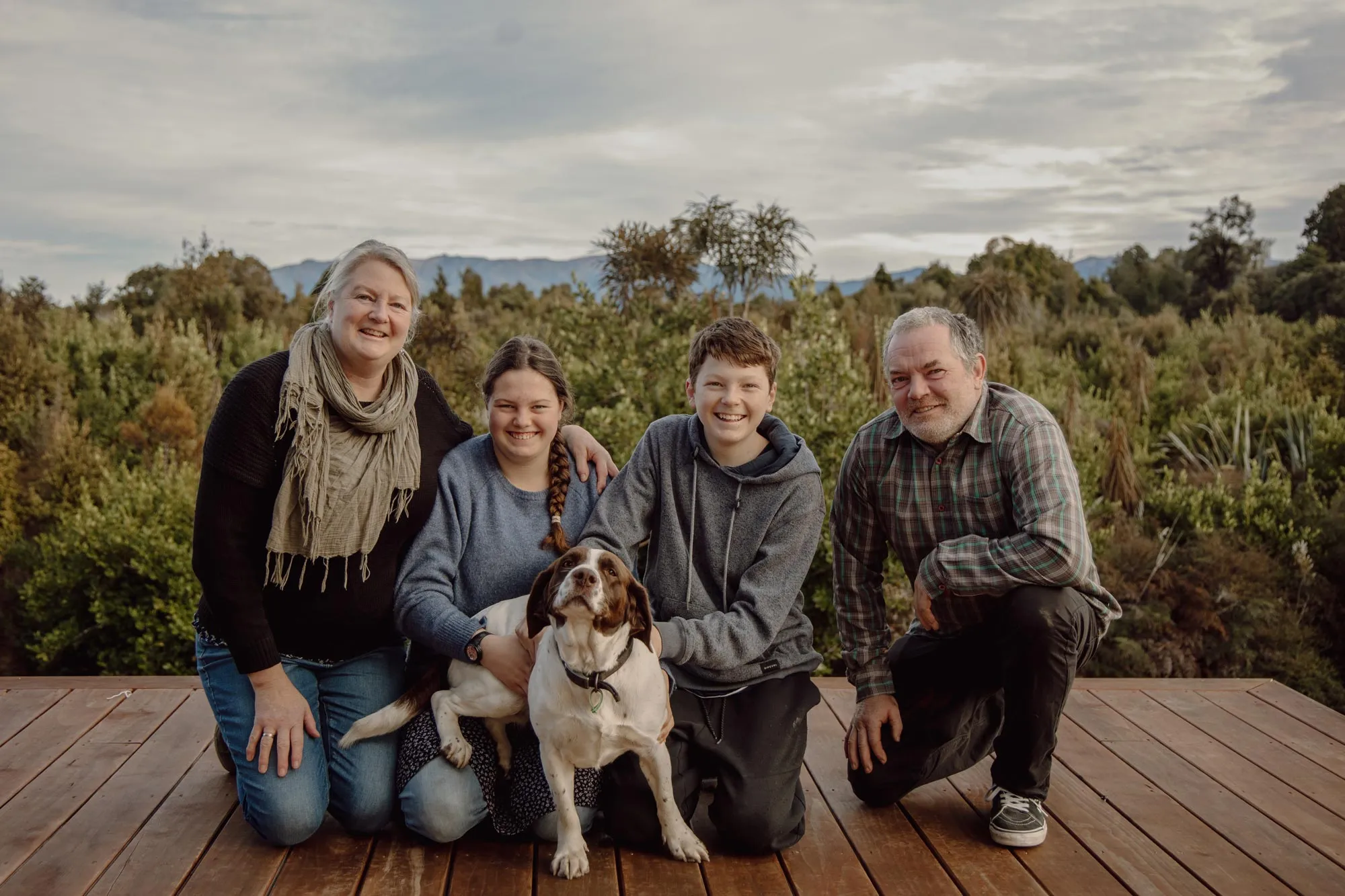 A family of four smiling and kneeling on a wooden deck outdoors with a dog in front of a green forest backdrop.