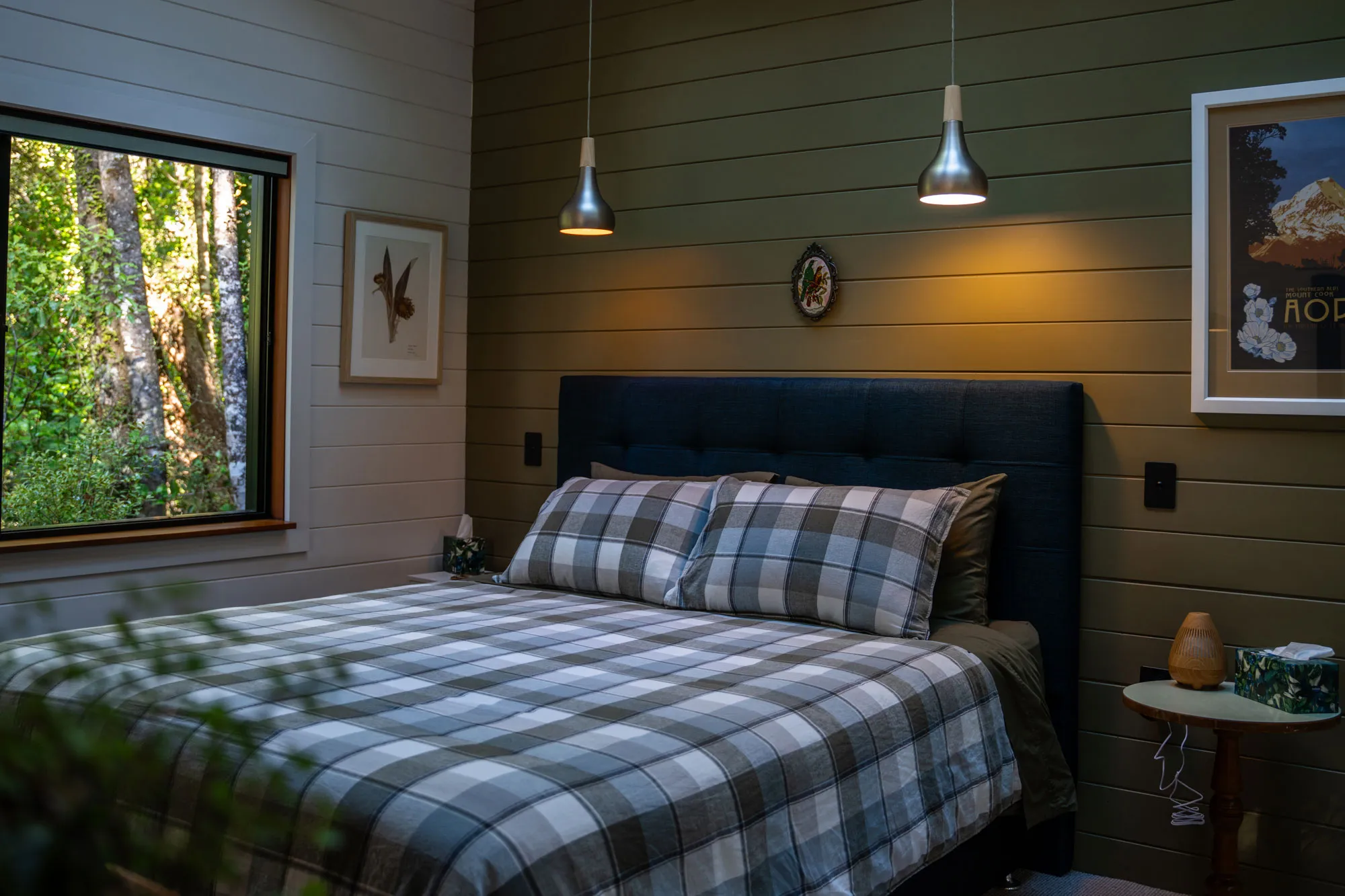 Cozy bedroom with a checkered bedspread and pillows, dark blue headboard, wooden walls, two pendant lights, framed artwork, and a window showing green trees outside.