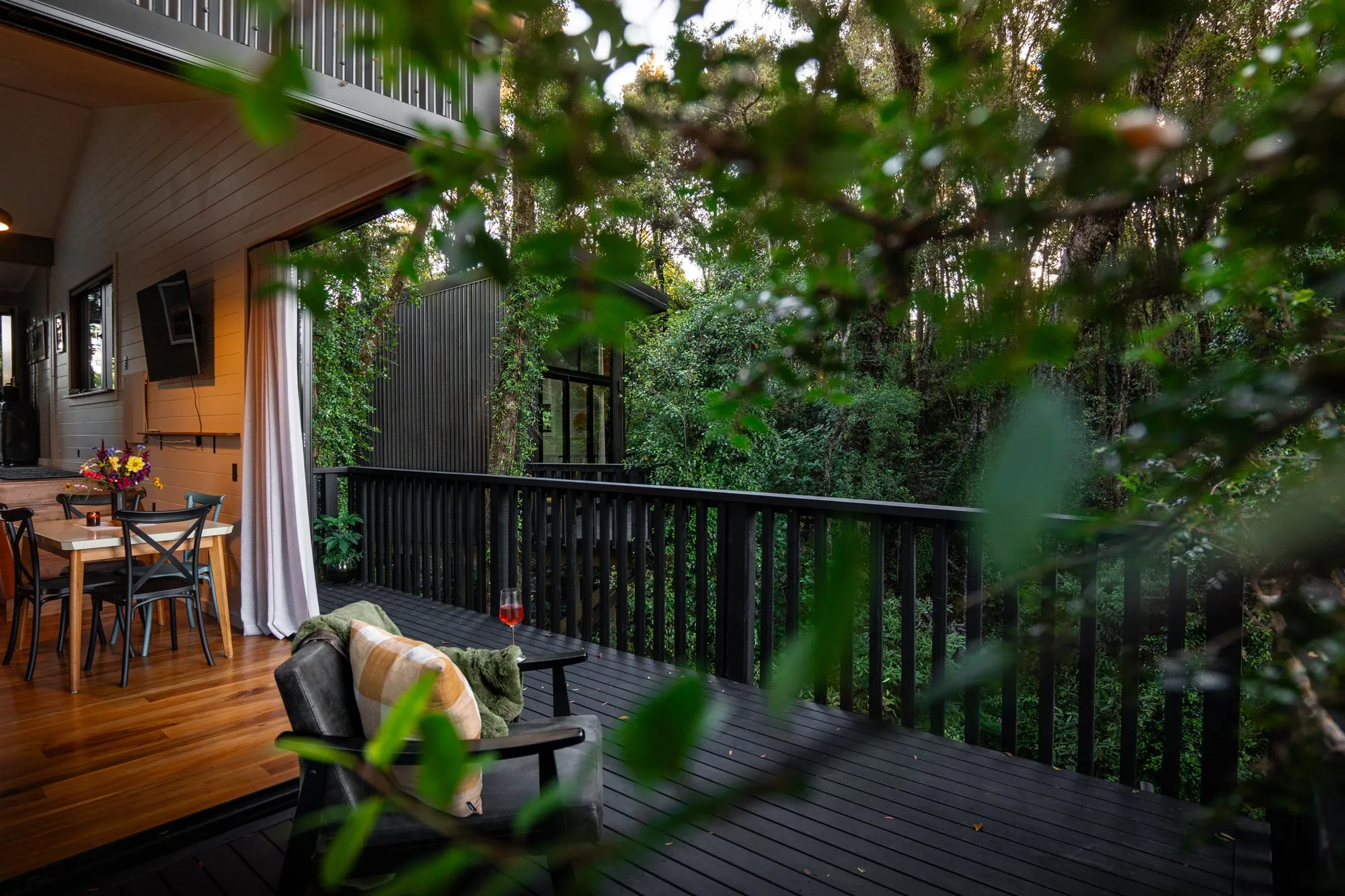Outdoor deck with a chair, plaid pillow, small table with a glass of red drink, and a dining table inside a house surrounded by lush green forest.