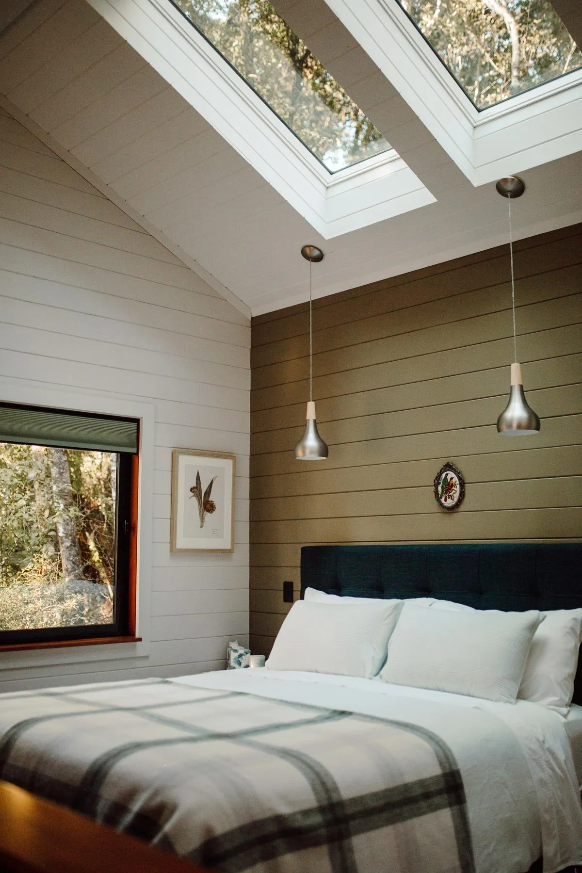 Cozy bedroom with a large bed, plaid blanket, two hanging pendant lights, skylights, and a window showing trees outside.