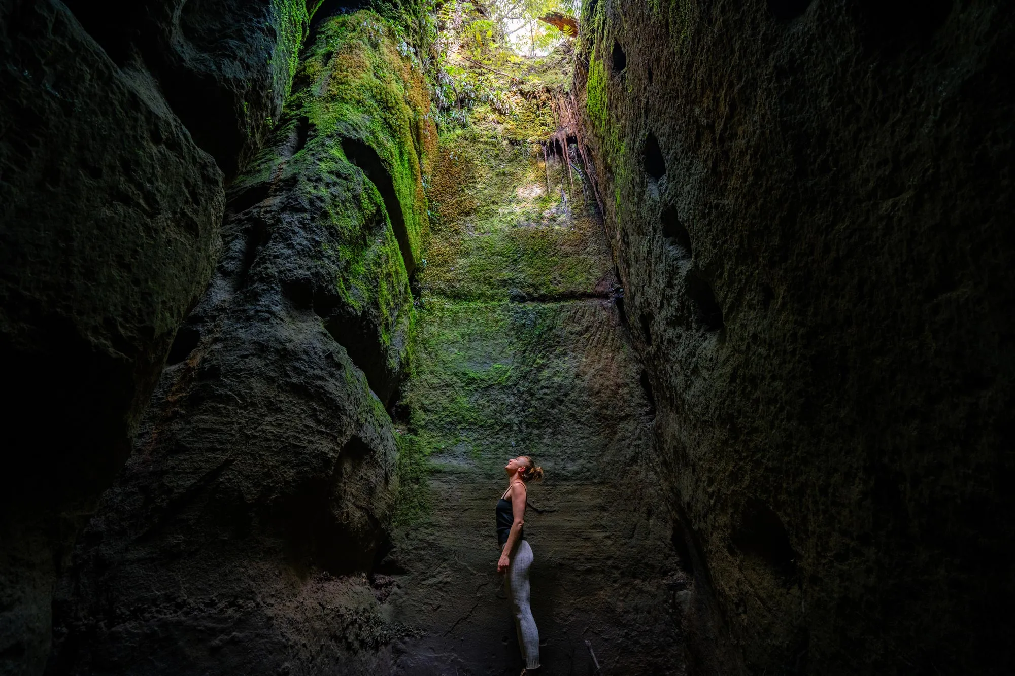 Woman standing inside a moss-covered, narrow rock crevice looking upward toward the light.