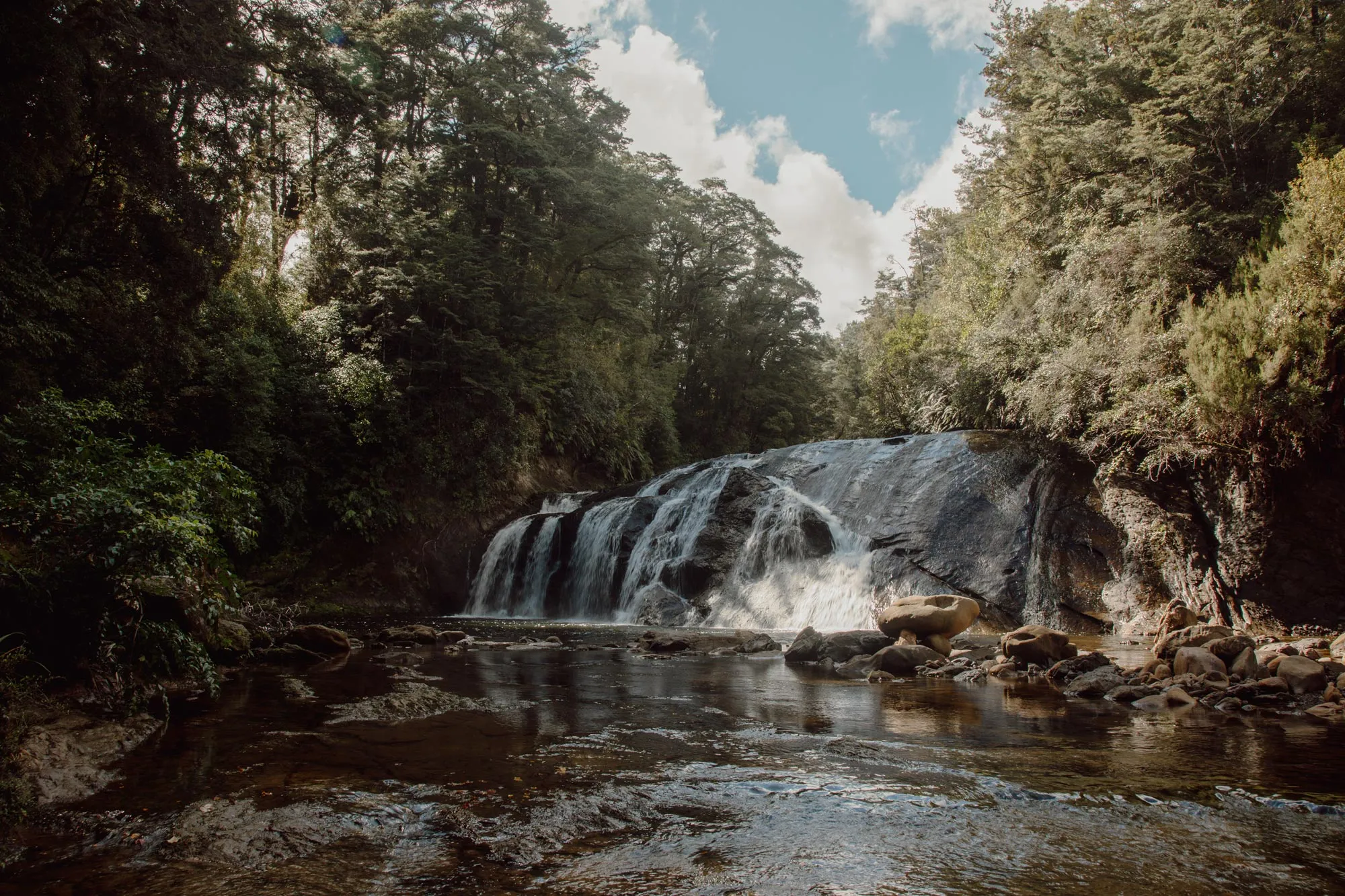 Small waterfall flowing over smooth rocks surrounded by dense green forest under a partly cloudy sky.