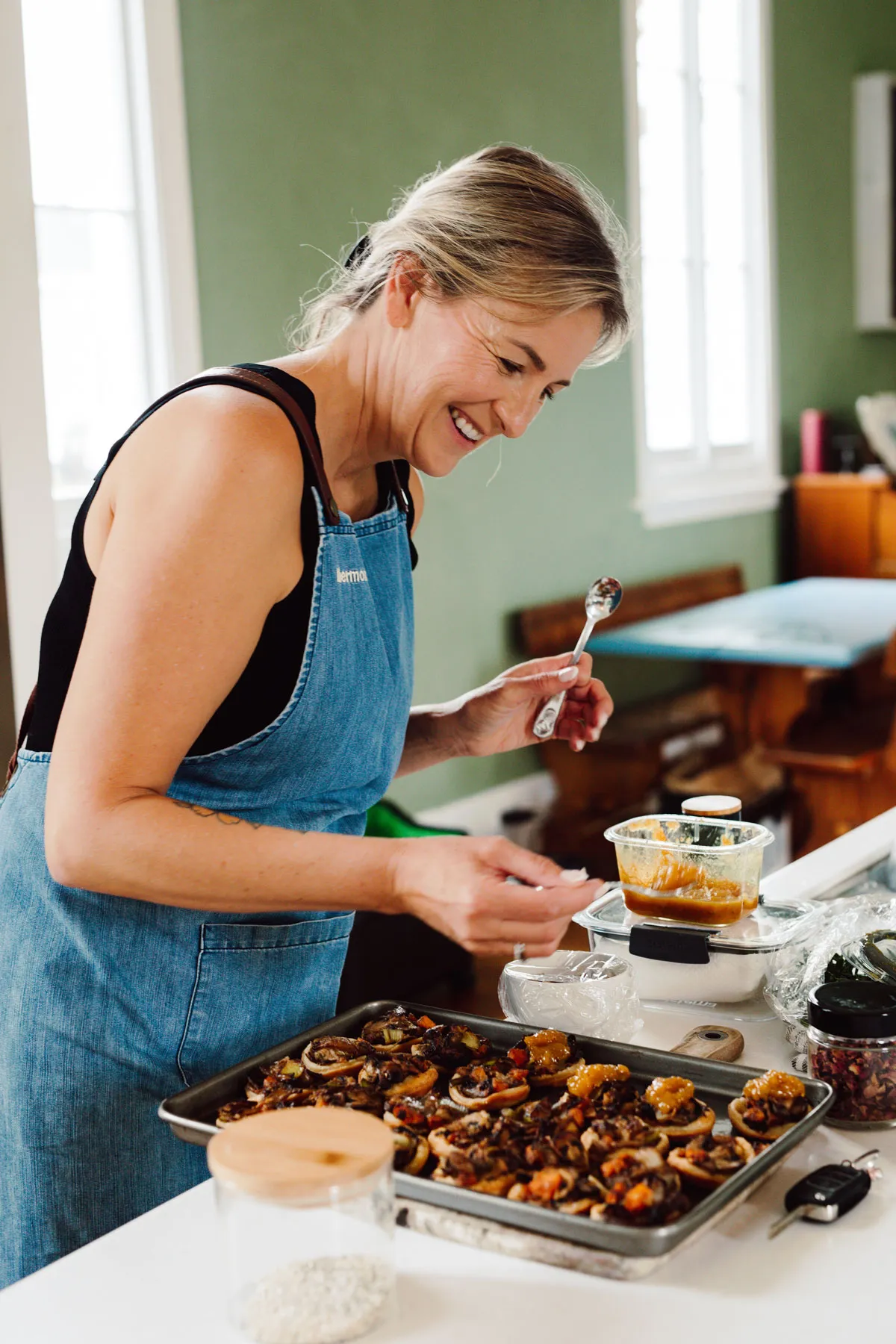 Woman in a blue apron spooning a topping onto baked mushrooms on a tray in a kitchen.