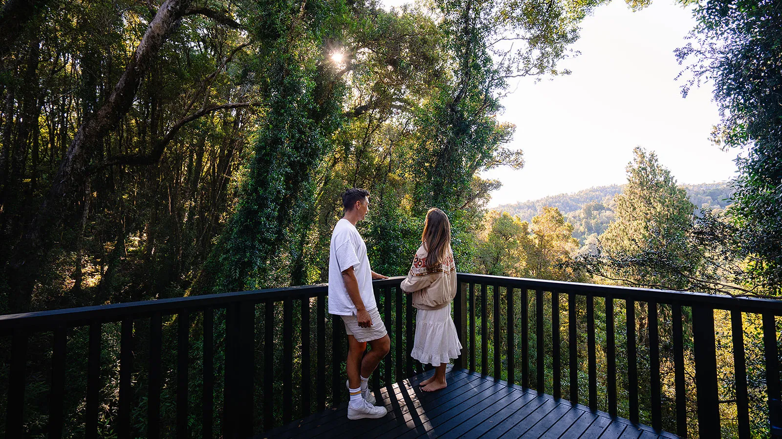 A man and woman standing on a wooden deck overlooking a sunlit forest and distant hills.