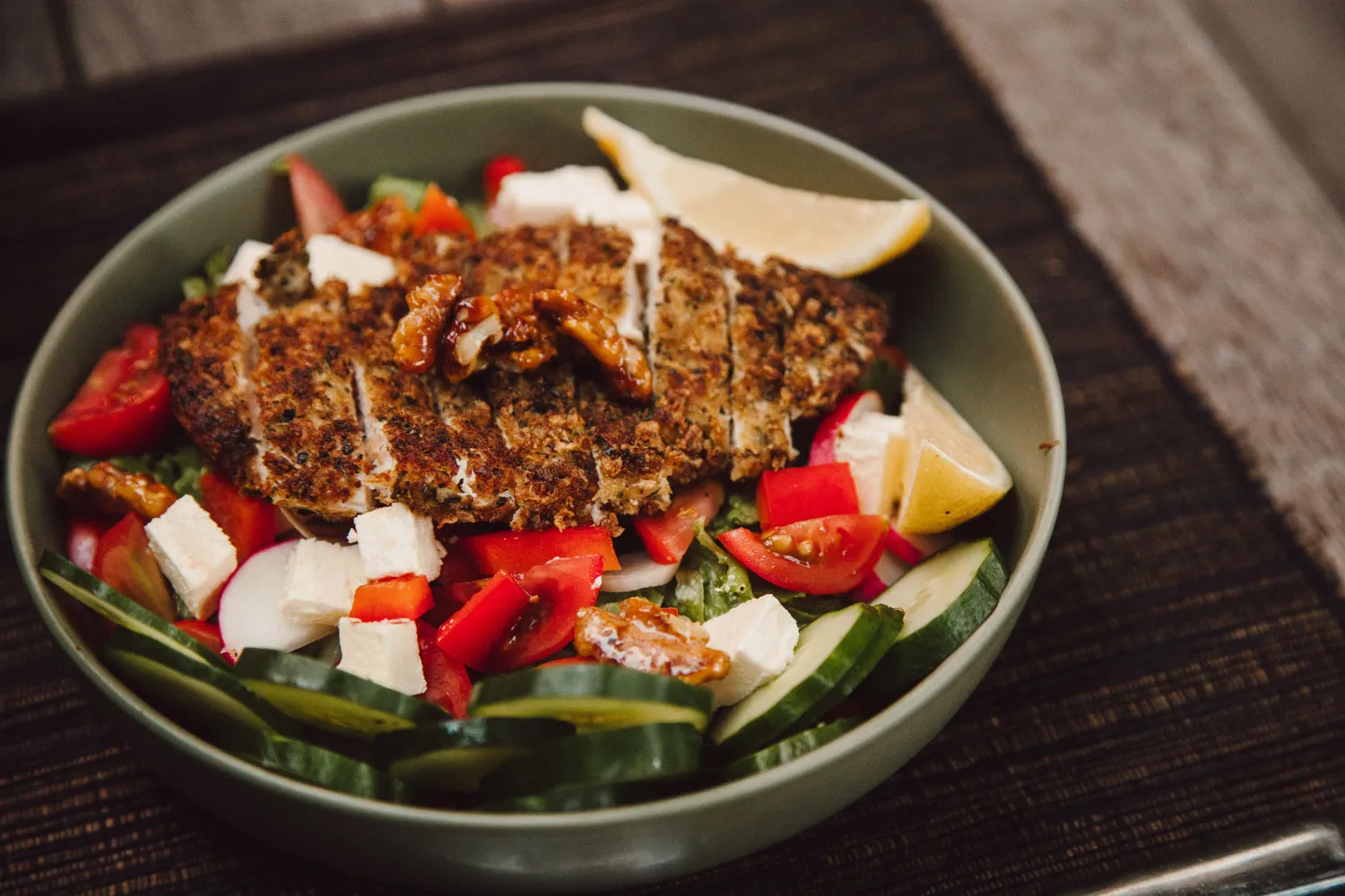 Bowl of salad with sliced grilled chicken, cucumber slices, cherry tomatoes, feta cheese cubes, walnuts, and lemon wedges on a dark placemat.