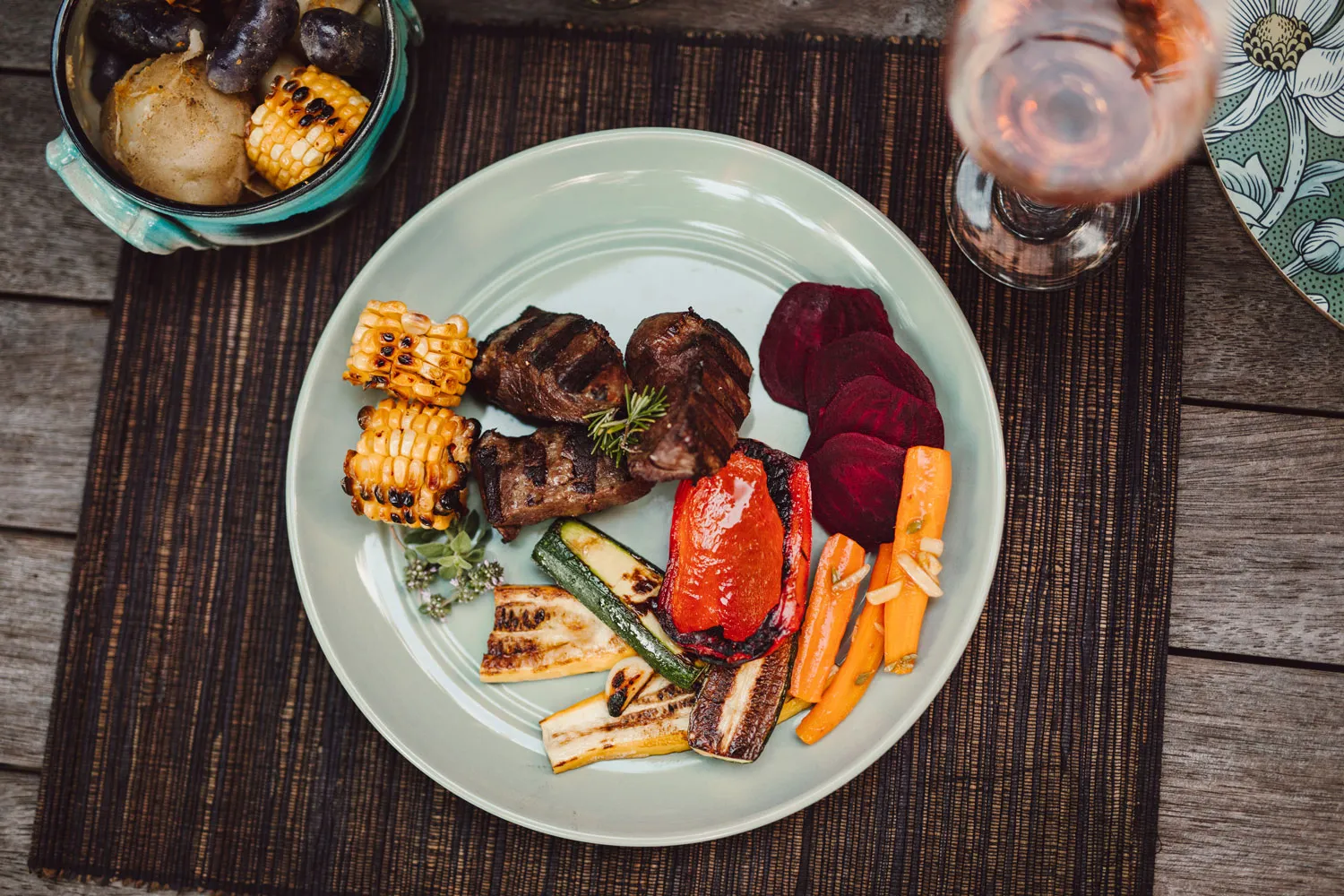 Plate with grilled beef, roasted corn, sliced beets, carrots with garlic, grilled zucchini and red bell pepper on a wooden table.