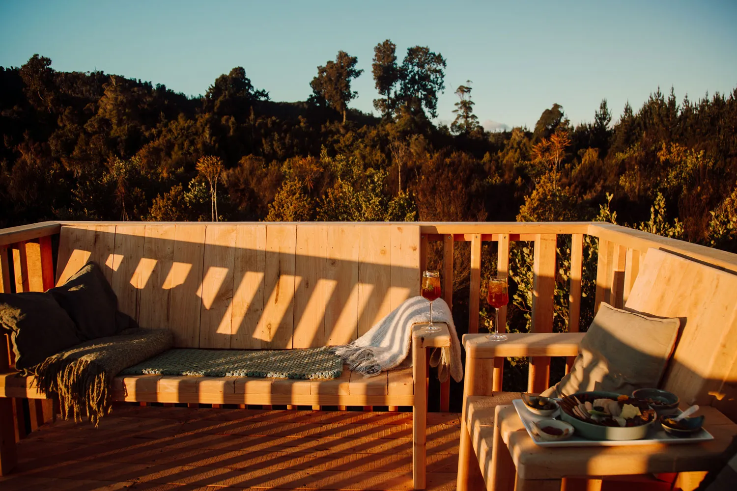 Wooden outdoor seating area with cushions, blankets, two glasses of iced drinks, and a tray of snacks overlooking a forest at sunset.