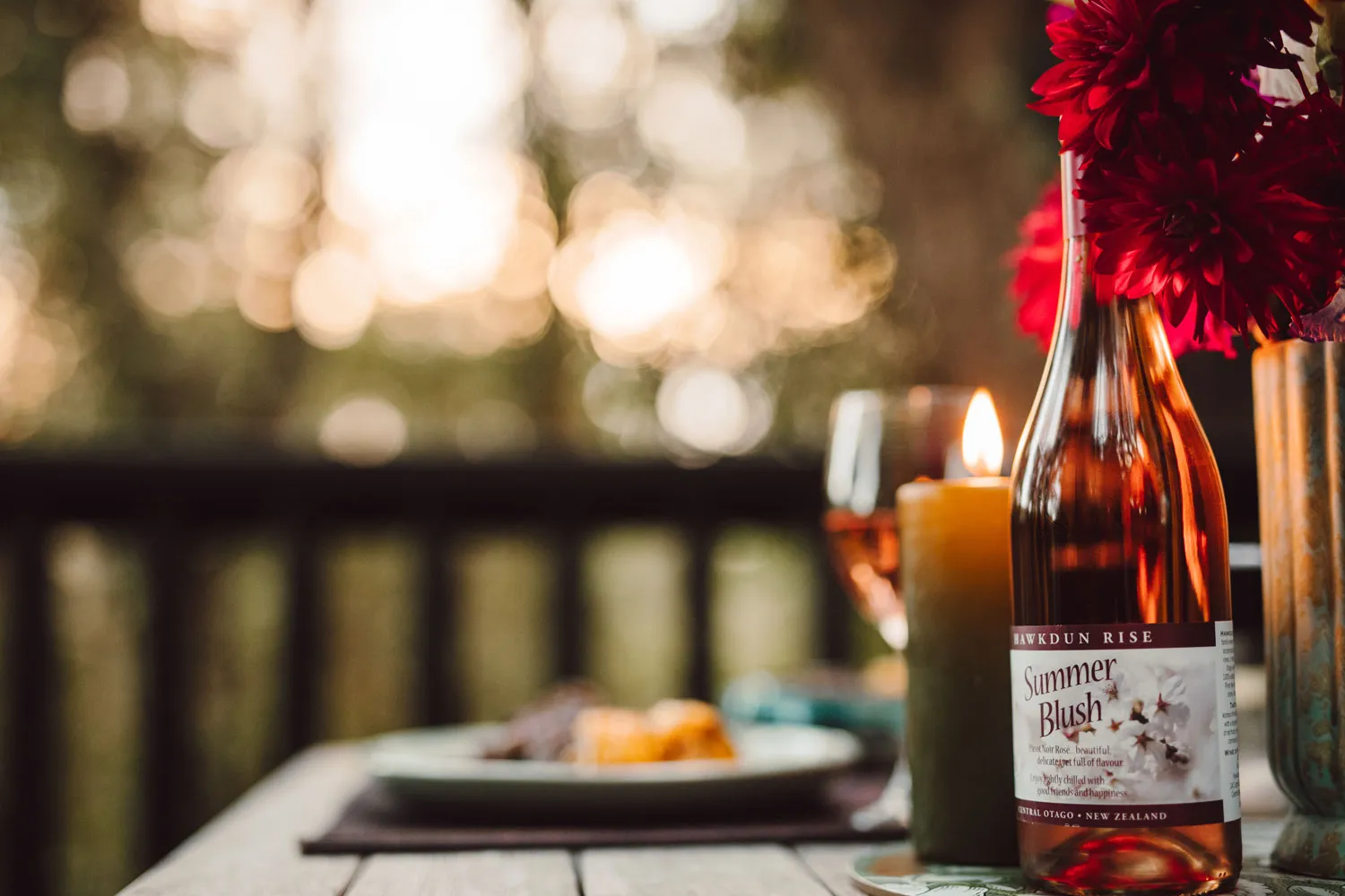 Bottle of Summer Blush rosé wine next to a lit candle and red flowers on a wooden table with a blurred outdoor background.