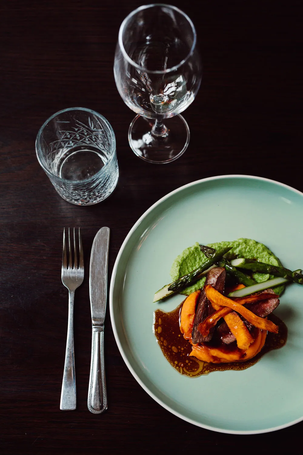 Gourmet dish with sliced meat, asparagus, carrot strips, and green puree on a pale green plate, accompanied by a knife, fork, empty water glass, and wine glass on a dark wooden table.