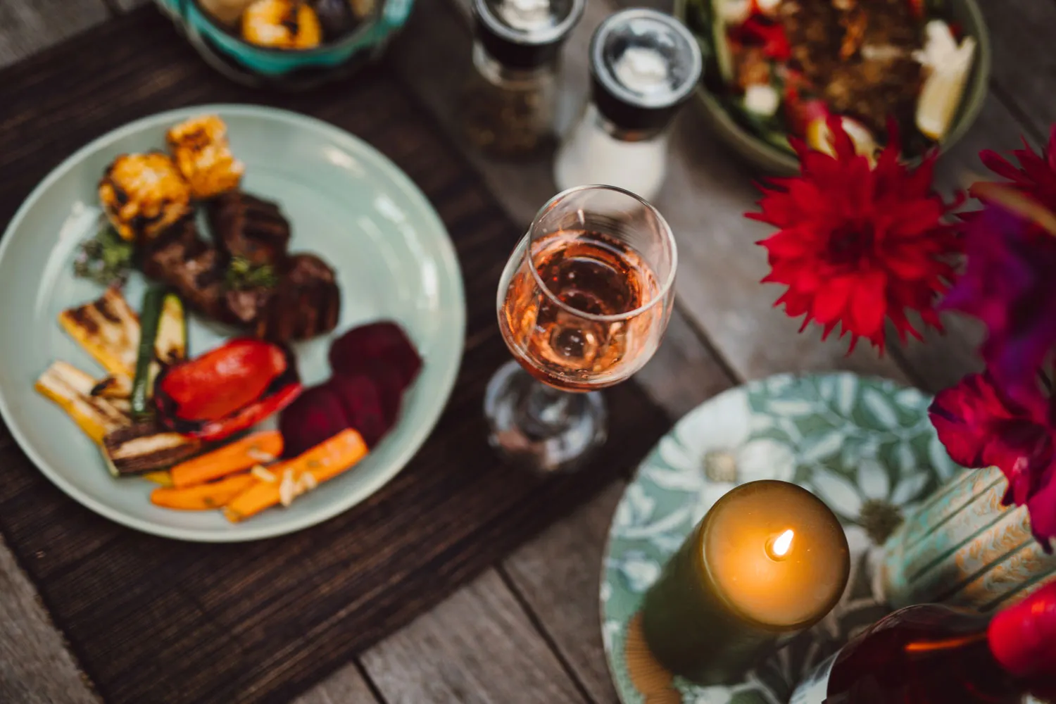 Table setting with a plate of grilled vegetables and meat, a glass of rosé wine, a lit candle, and red flowers.