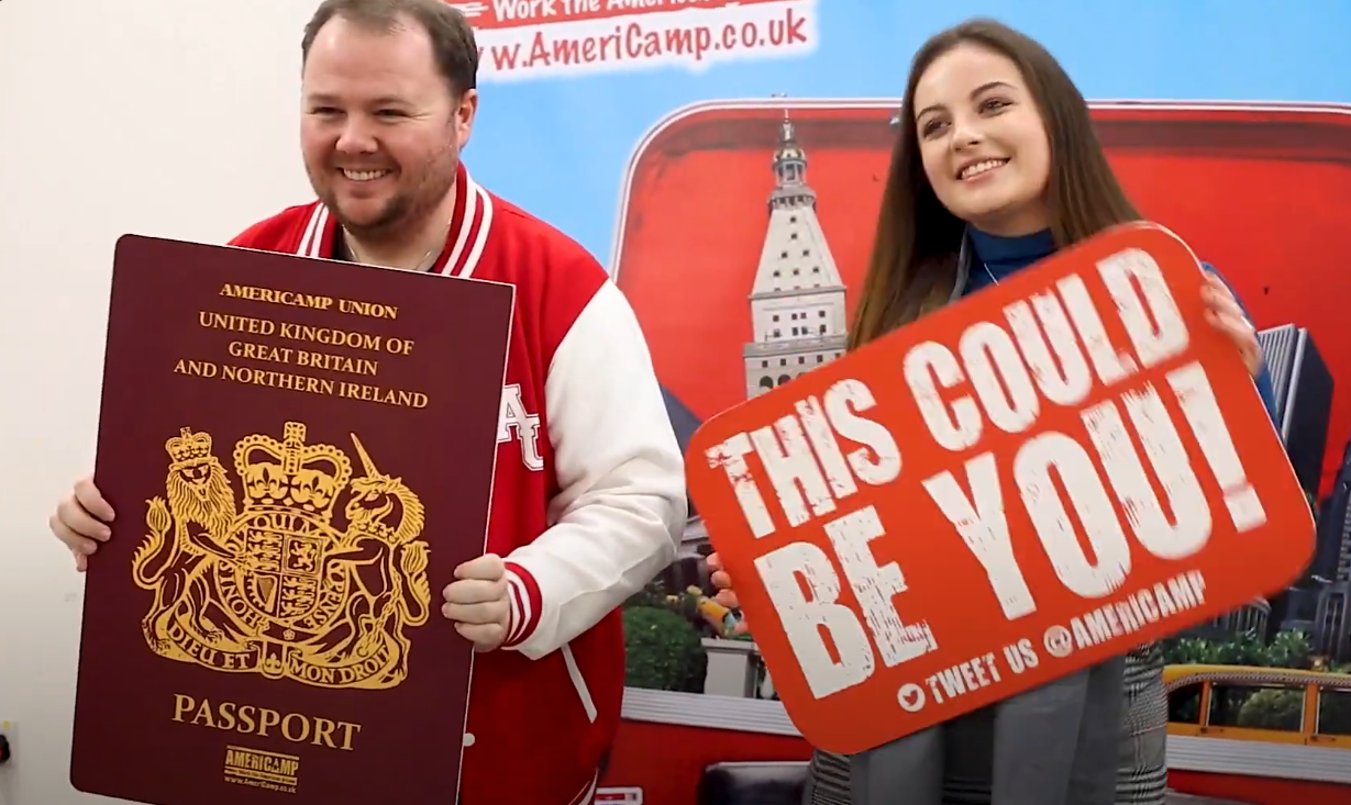 Two members of AmeriCamp pose for an image in front of AmeriCamp's UK banner, holding large novelty passports and placards that say "This could be you." 