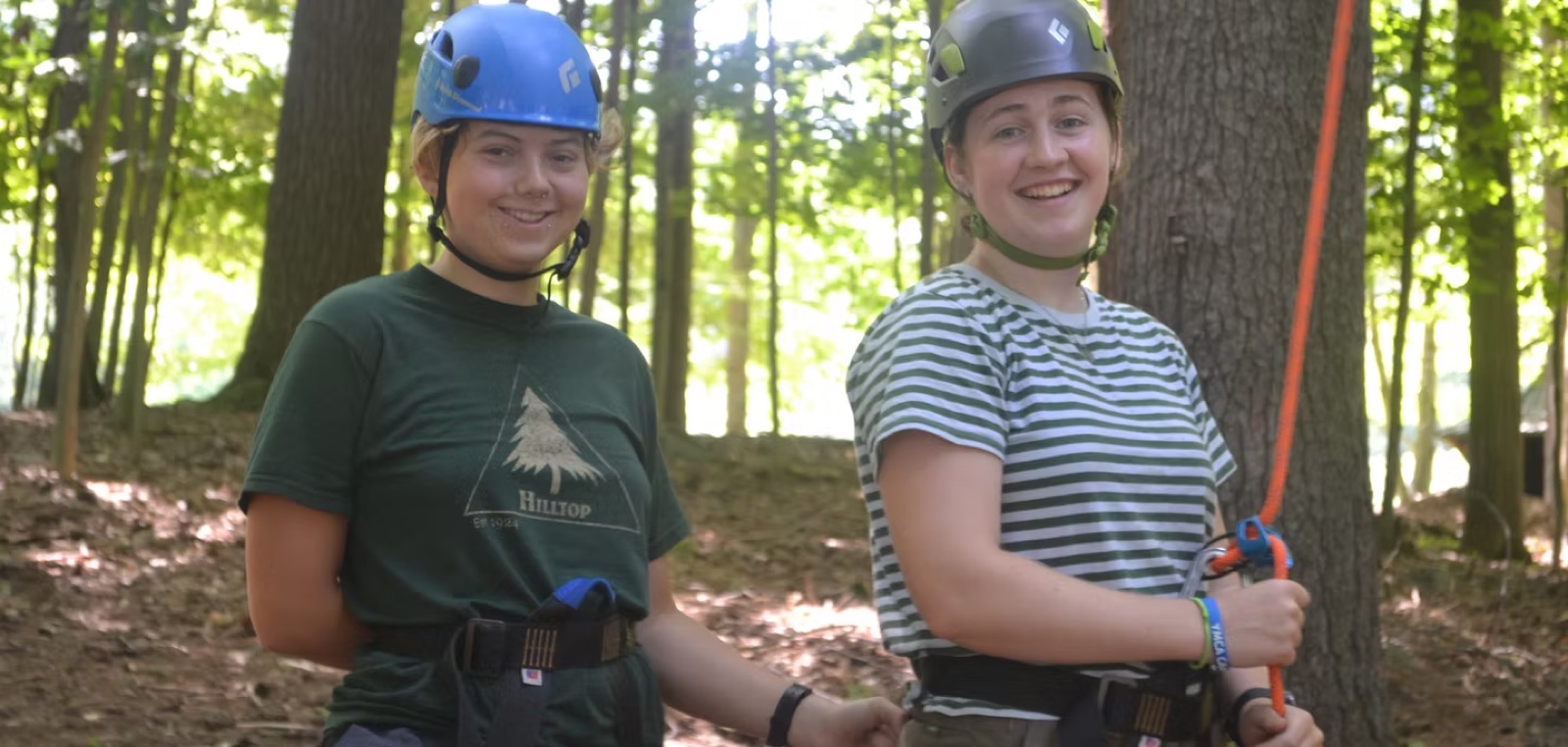 Two camp staff in climbing helmets and harnesses at the ropes course in the woods at YMCA Camp Chingachgook