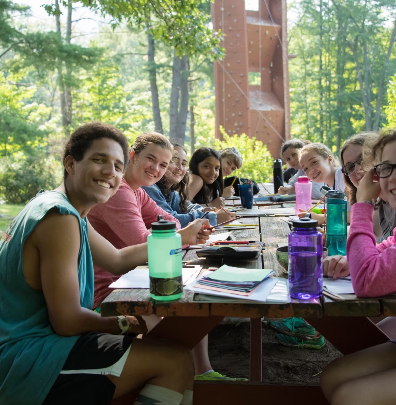 A camp counsellor and campers smiling at a picnic table beside the climbing tower at YMCA Camp Chingachgook
