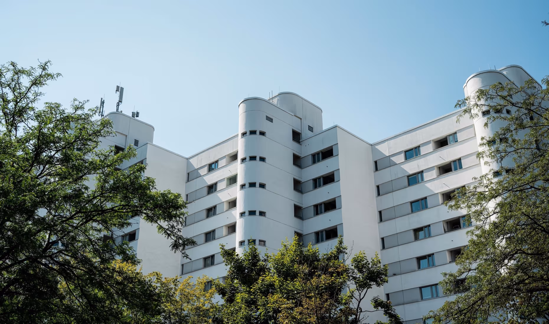 Modernes weisses Wohngebäude mit abgerundeten Ecken und mehreren Stockwerken, im Vordergrund grüne Bäume und blauer Himmel im Hintergrund.