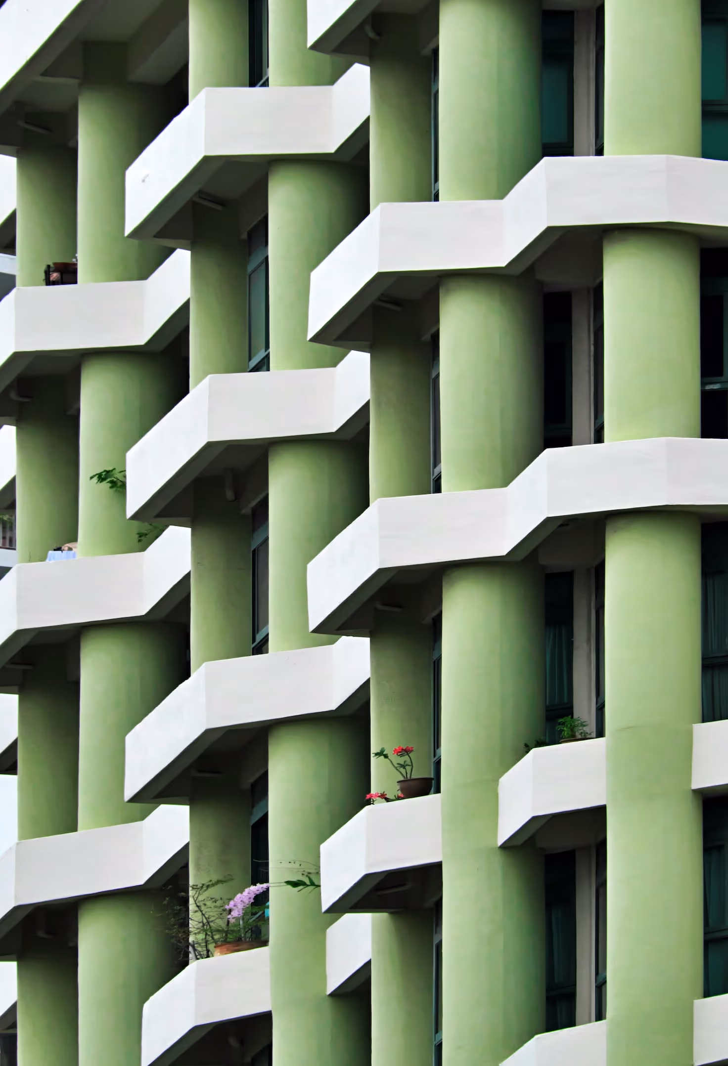 Fassade eines grünen Wohnhauses mit weissen, versetzt angeordneten Balkonen und runden Säulen, die ein rhythmisches Muster bilden.