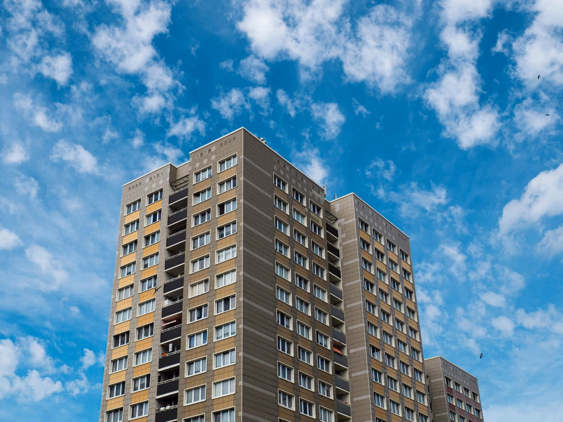 Hohes Wohngebäude mit braun-beiger Fassade und vielen Fenstern, fotografiert vor einem blauen Himmel mit weissen Wolken.