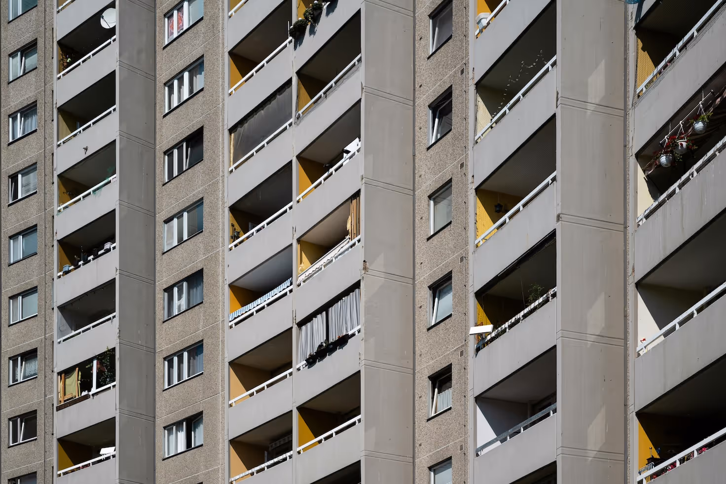 Nahaufnahme einer Plattenbau-Fassade mit vielen offenen Balkonen, Geländer und Wänden in Grau und Gelb, teilweise mit Vorhängen und Hängepflanzen.