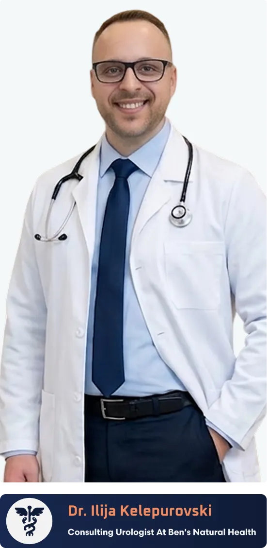 Smiling male doctor wearing glasses, a white coat with a stethoscope, a blue shirt, and tie standing in a hallway.